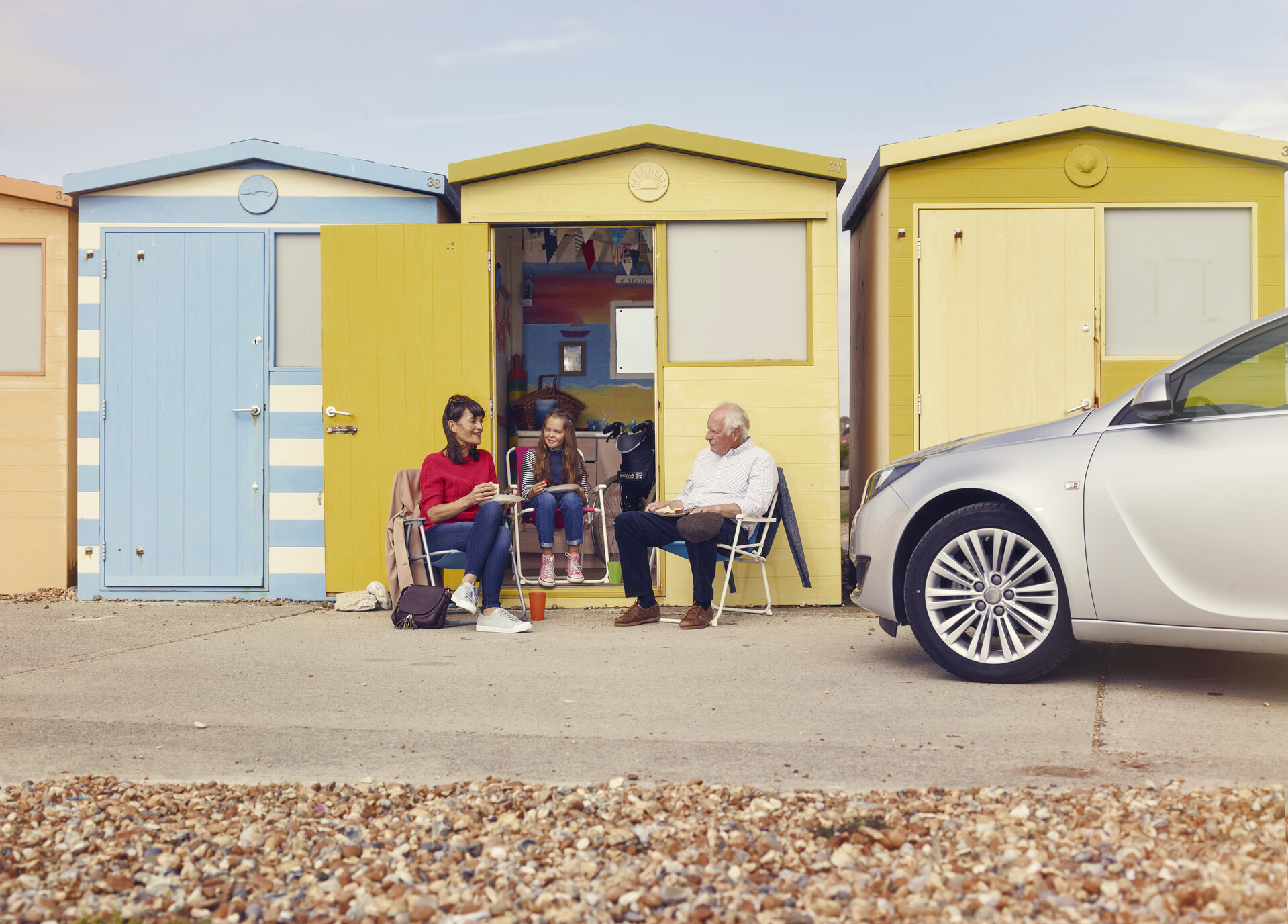 An older man, a woman and a young girl sit outside a yellow beach hut, next to a car