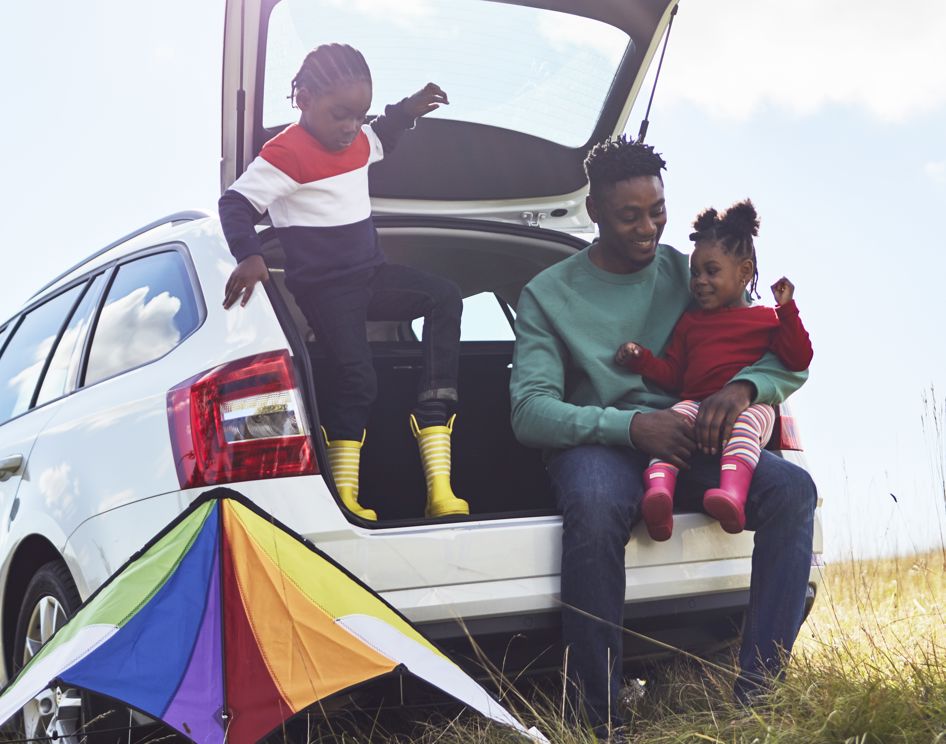 Man and two children sitting in the open boot of a car, with a kite in a field