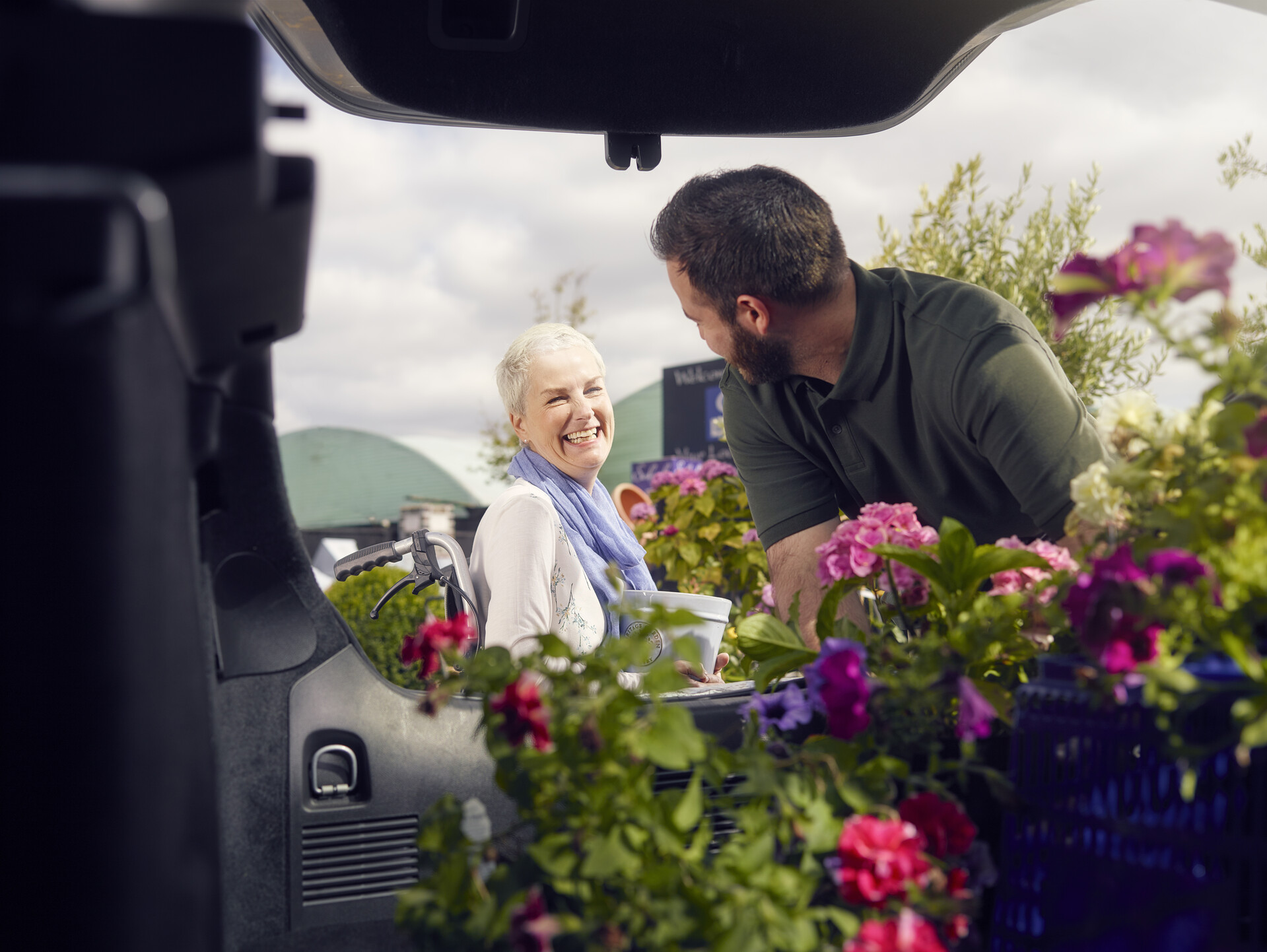 An older woman smiling and talking to a younger man, while he loads flowers into his boot