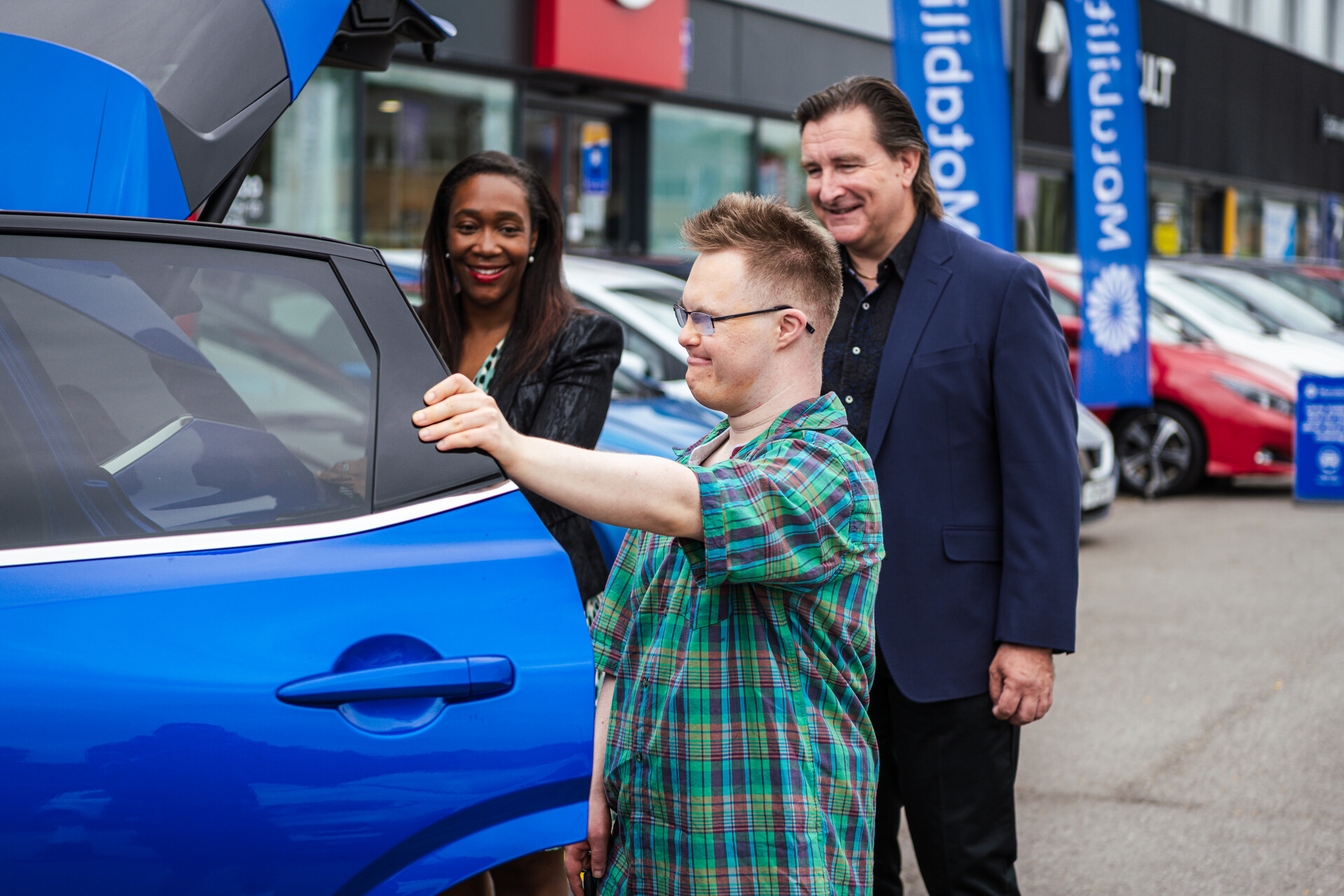 A young man looks inside a car at a dealer, with a smiling man and woman