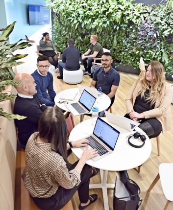 A big group of colleagues sit in two groups and talk, in an open office area