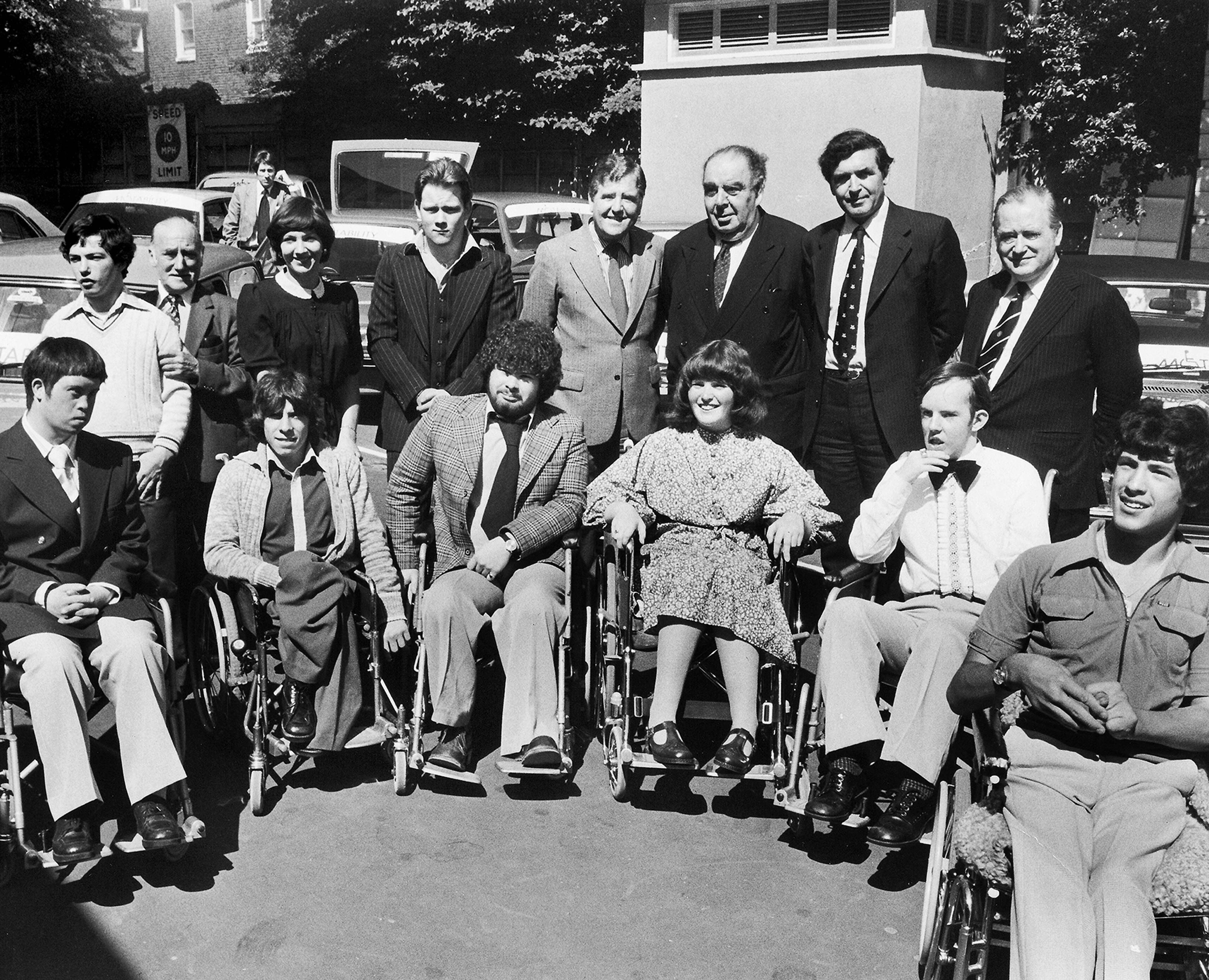 Six young people in wheelchairs pose for a black-and-white photo, with car dealers and Motability Operations staff