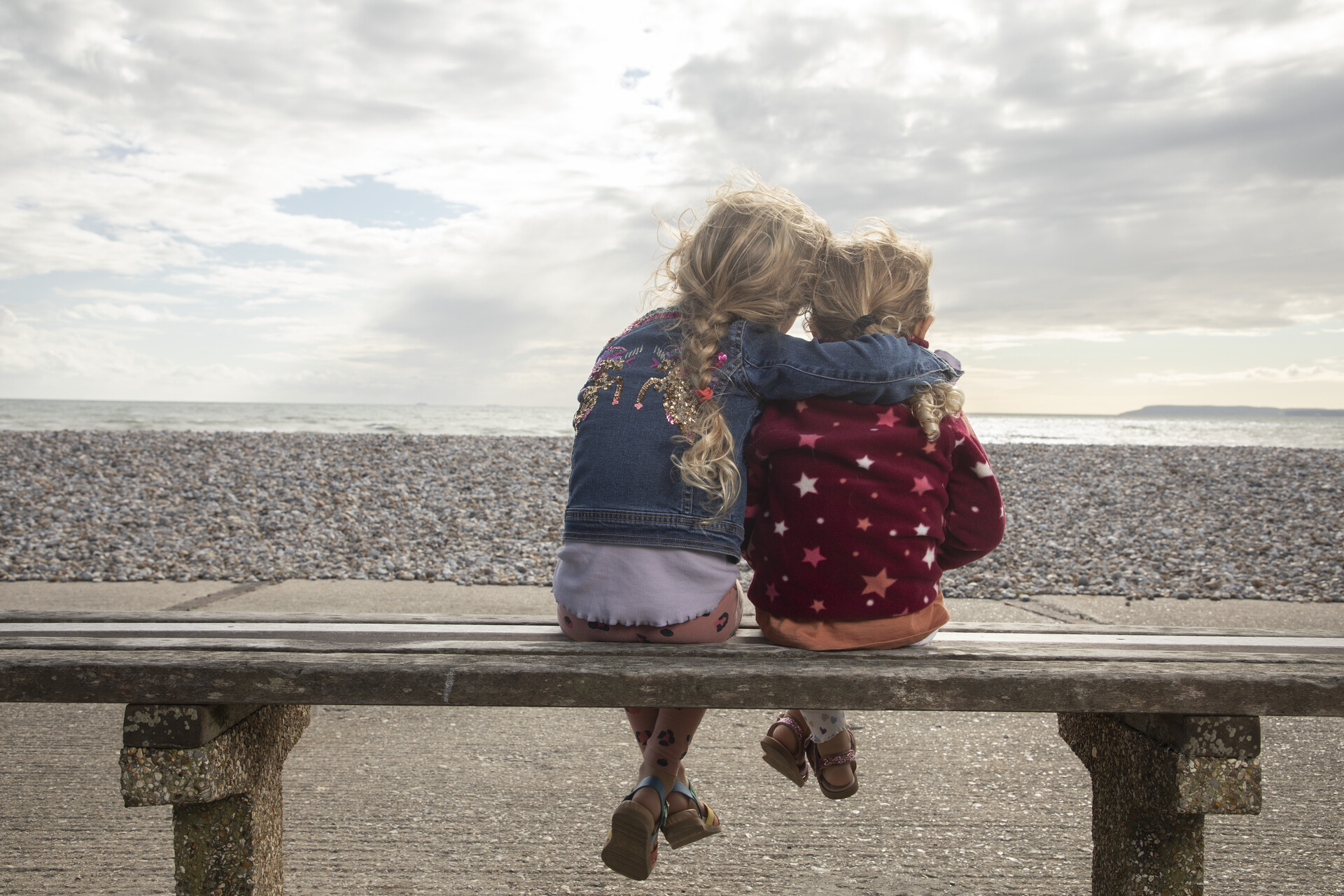 Two young girls sitting on a bench and hugging, looking at a beach