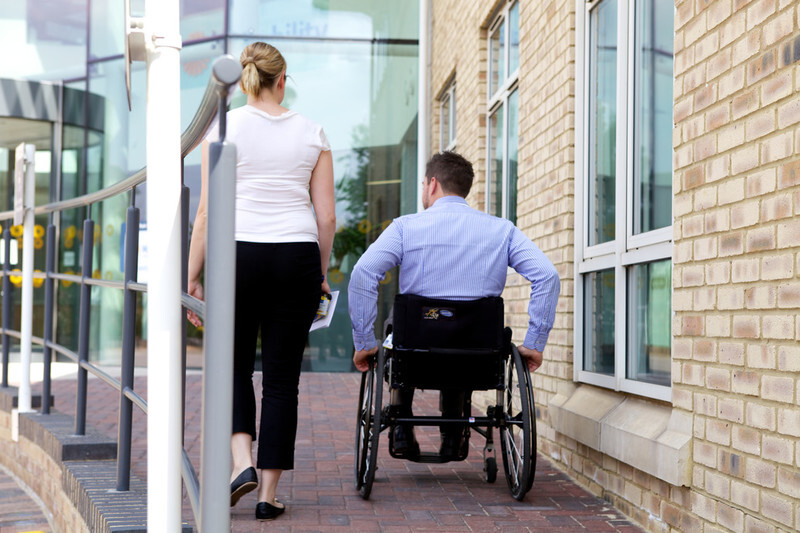 Woman walks up ramp next to man in wheelchair, photographed from behind