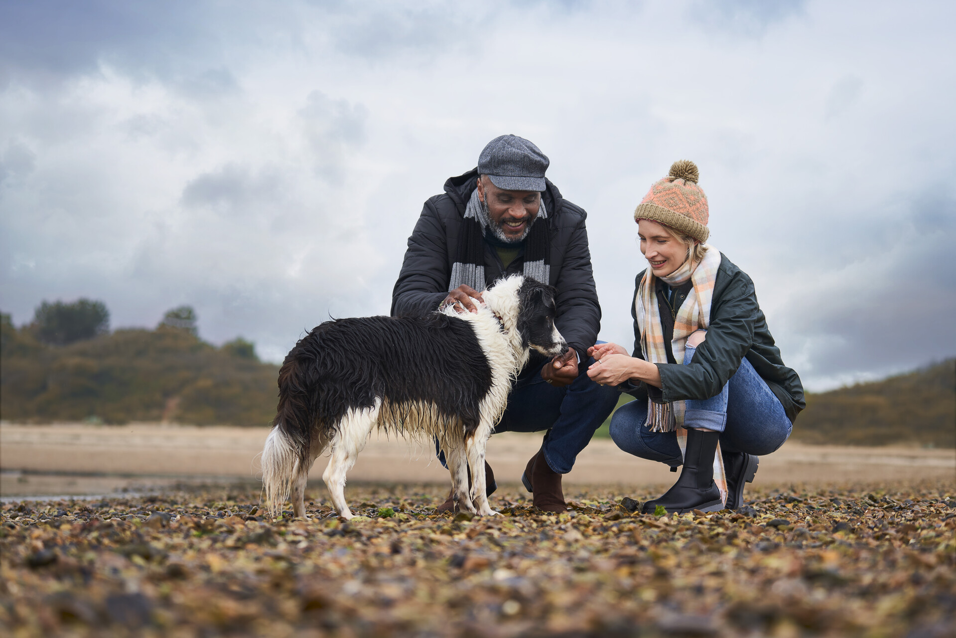 A man and a woman petting a dog on a pebble beach