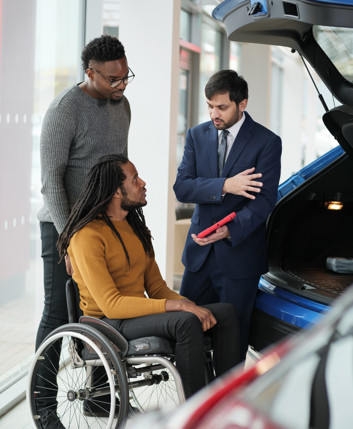 A dealer shows a car boot to a man in a wheelchair and man standing behind him