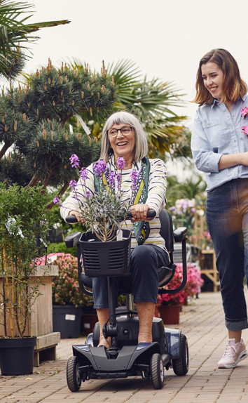 A smiling older woman on a mobility scooter, next to a smiling woman holding flowers