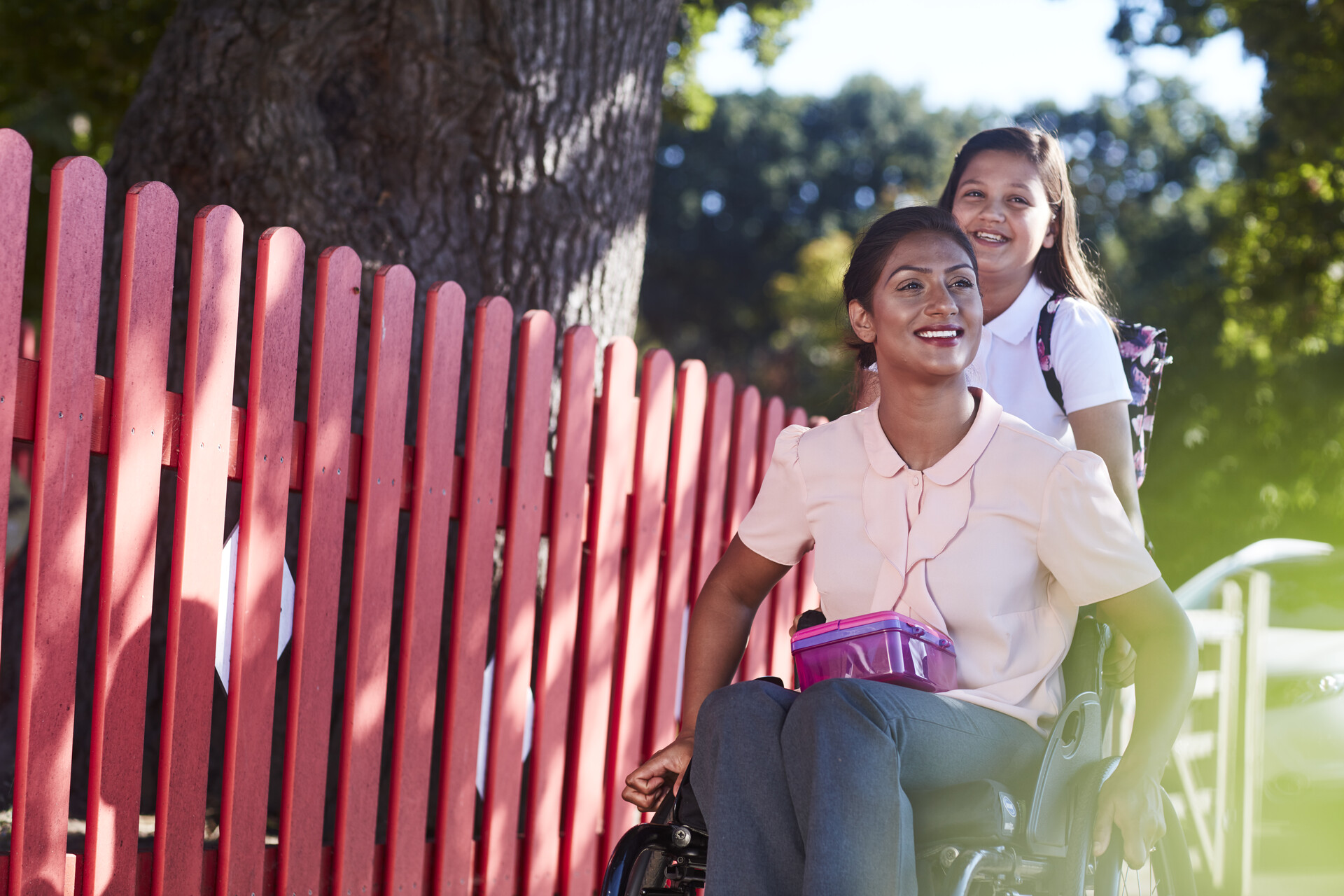 A smiling woman in a wheelchair with a lunch box, and a schoolgirl following behind her