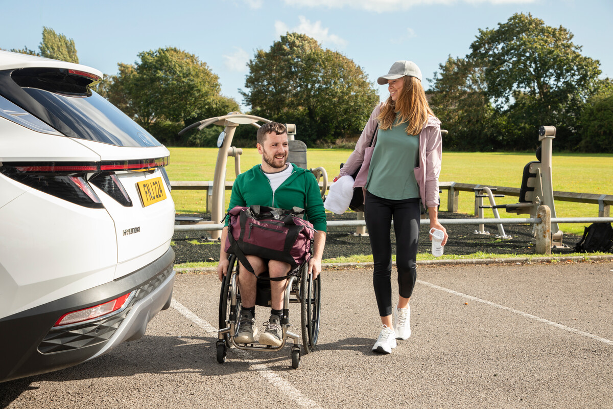 A woman walking with a man in a wheelchair, wearing sports gear, next to a car
