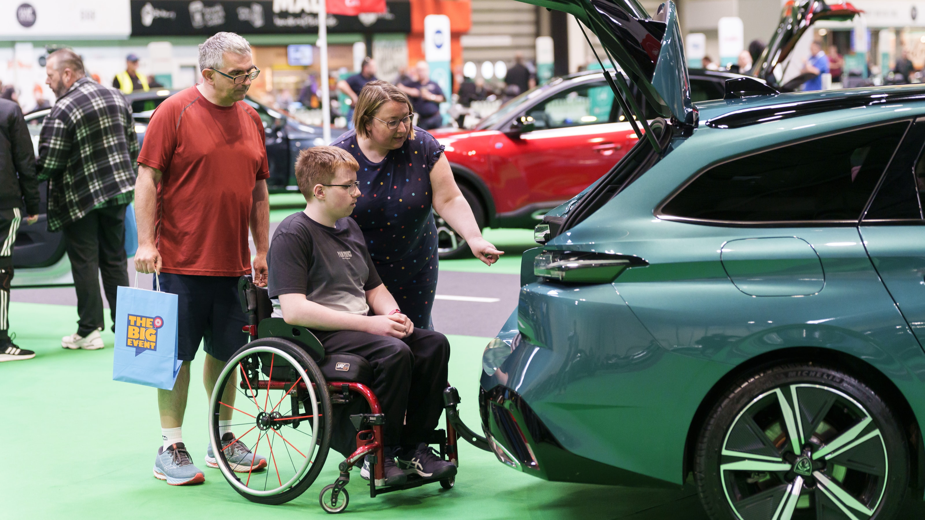 A family of three look in the boot of a car. The sits in a wheelchair and his parents stand either side of him.